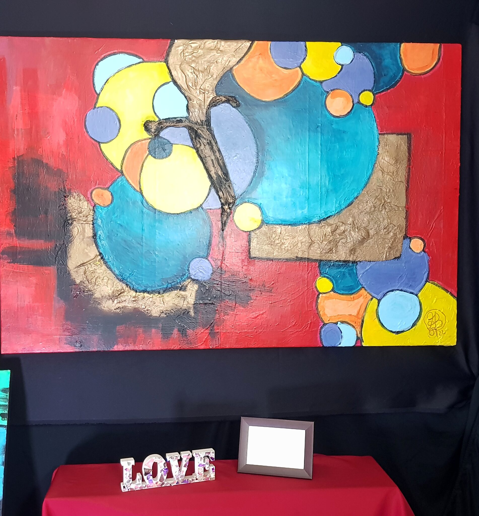 Fotografie eines Kunstwerkes etwa 1 m entfernt aufgenommen auf schwarzem Hintergrund das Werk ist groß hat einen roten Hintergrund und gelbe orange blaue und lilane Reise in verschiedenen Größen im Vordergrund es hat zwei Flächen die 3D mäßig aus dem Berg kommen diese sind Gold und Schwarz schattiert